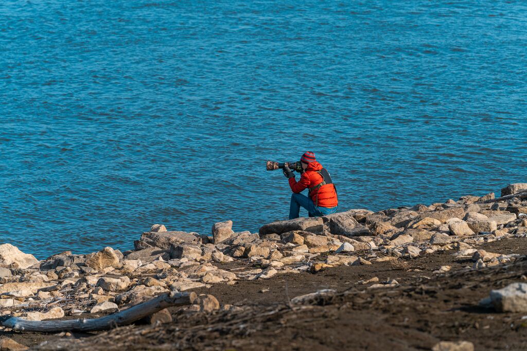 A person in a red jacket crouches at river's edge with a telephoto lens.