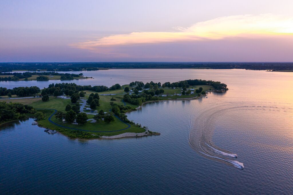 The sun sets on the lake and the sky has a pink afterglow. In the distance, there is a town spanning the banks. There is a cape that extends out into the water, and a community with homes and paved roads line the banks. There are two wave runners zipping through the water.