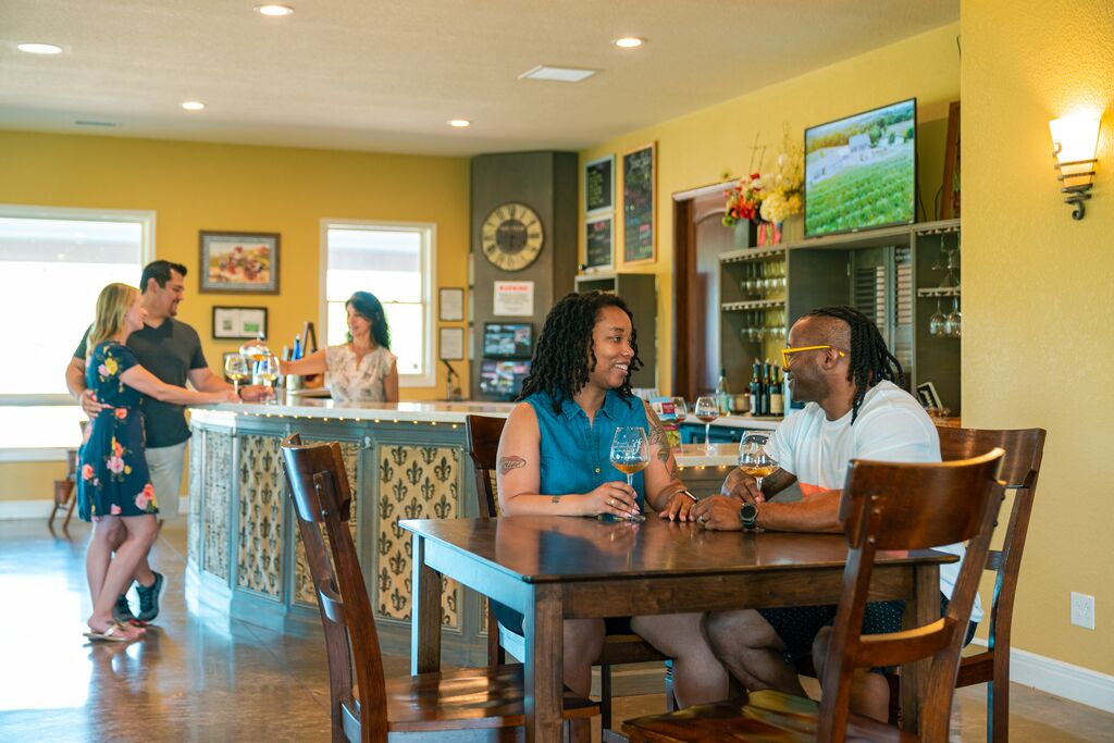 A couple sits at a table in a wine tasting room sipping on white wine while another couple is served wine at a bar in the background