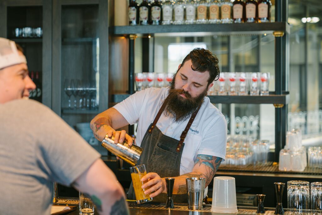 A bartender pours a cocktail into a clear glass. The customer is laughing and talking to someone. Whiskey bottles line the shelves behind the bar.