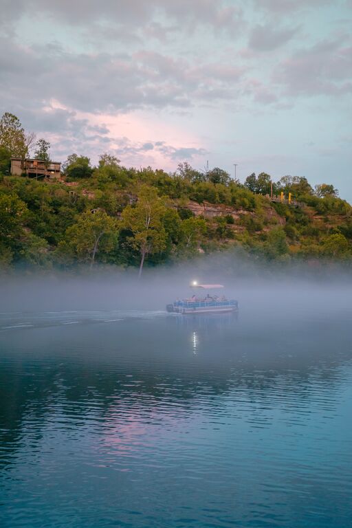 The sky is light blue and pink as the sun rises. There is a large bluff with trees covering most of it and a house on the bluff. Below, the blue water ripples and fog covers the lake. In the fog, there is a small motorboat.