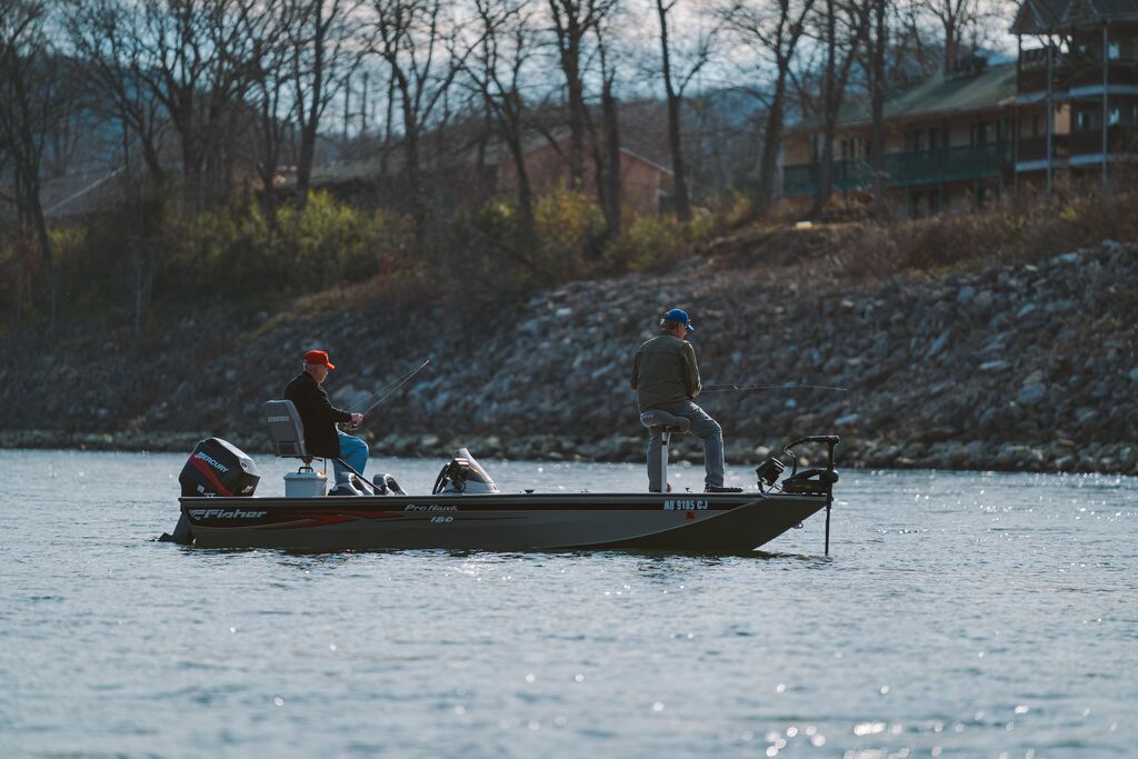 Two people fishing from a boat along a lake bank.
