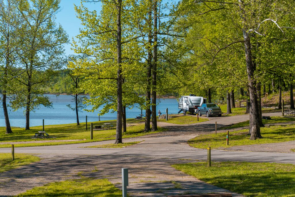 There are paved roads weaving throughout tall green trees. There is a blue lake at the edge of the bank, behind the trees and paved road. There is a black, large truck attached to an RV, parked beside the water. There are picnic tables and grills scattered throughout the area.