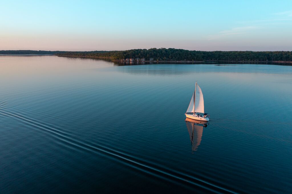 A sailboat sits on glass like lake with a sunset in the distance.